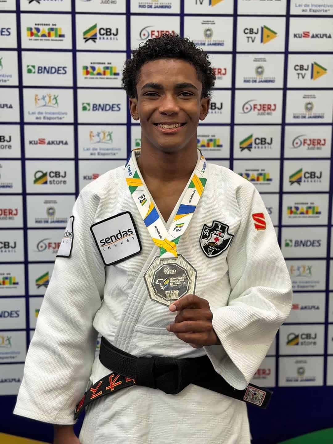 Atleta de judô com uniforme branco e cinturão preto, exibindo medalha e troféu, comemorando conquista na competição.
