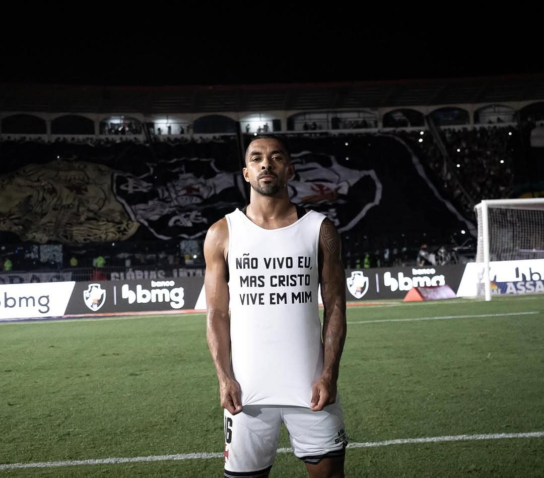 Texto alternativo: Jogador do Vasco vestindo uniforme branco, de pé no estádio à noite, com arquibancadas e torcida ao fundo.