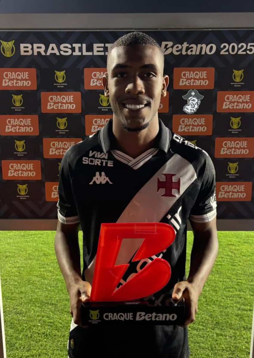 2. Alt: Jogador do Vasco segurando troféu na celebração da vitória, usando uniforme preto e branco, em estádio com painel de patrocinadores ao fundo.