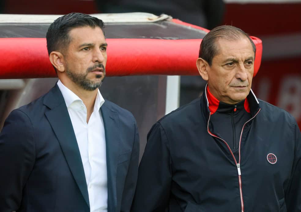Homem de terno e homem de casaco preto em evento esportivo.