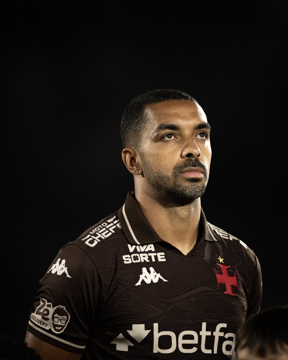 Jogador do Vasco em retrato, uniforme com logos de patrocinadores, fundo preto, expressão séria.
