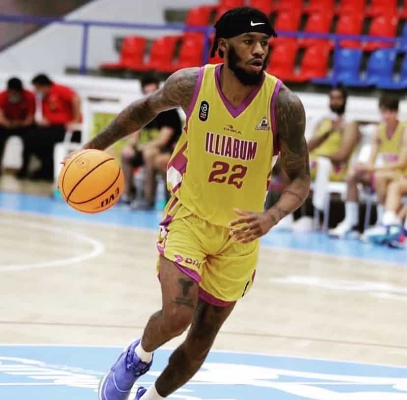 Atleta do Vasco com uniforme amarelo e roxo, driblando durante jogo de basquete. Fundo com colegas e torcida no ginásio.