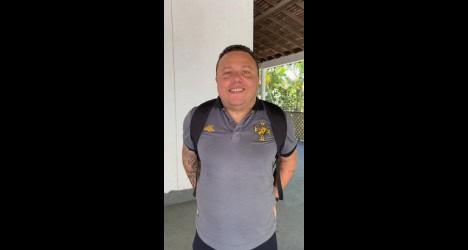 Homem sorrindo com camisa do Vasco e mochila preta, em ambiente aberto com paredes brancas e vegetação ao fundo.