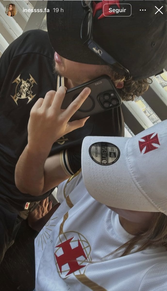 Imagem de um torcedor do Vasco usando capuz e camisa do clube, segurando um celular perto do rosto, em ambiente interno. A camisa apresenta o escudo do Vasco da Gama, e o torcedor parece estar concentrado na tela.