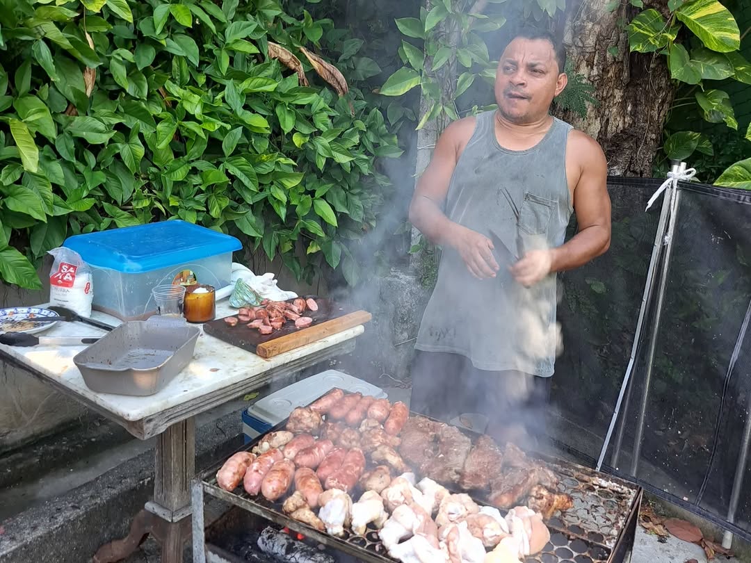 20251130-142351-1-7 Churrasco em churrasqueira ao ar livre, carnes assando na grelha oriental, cercado por vegetação verde ao fundo.