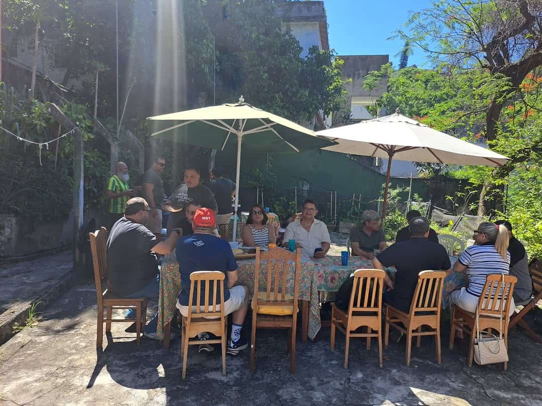 20251130-142352-1-8 Pessoas ao redor de mesa ao ar livre com guarda-sóis brancos, em ambiente ensolarado e arborizado, participando de encontro social.