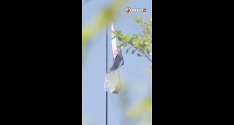 Imagem de uma bandeira do Vasco da Gama ao vento em um dia ensolarado, pendurada em um cabo, com céu azul ao fundo e algumas folhas verdes ao redor.