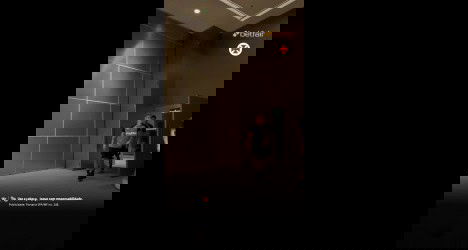 Homem com camisa cinza e short preto entrando no aeroporto, com outro ao lado, em fundo de parede escura e iluminação suave.