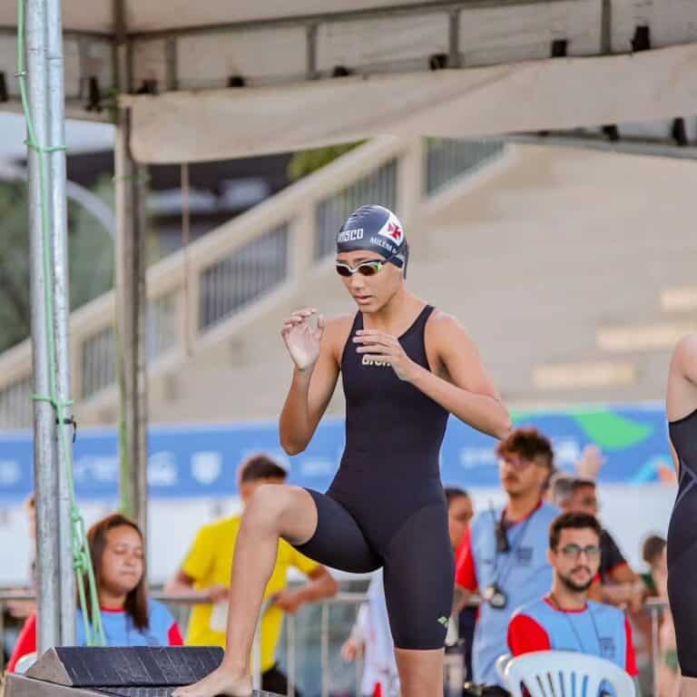 Texto alternativo: Nadadora do Vasco treinando na piscina, usando touca, óculos de proteção e traje de natação, com outros competidores ao fundo.