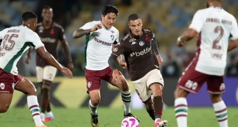 Imagem de jogadores do Vasco em ação durante partida, com um adversário, em campo de futebol, concentrados na jogada, com estádio ao fundo.