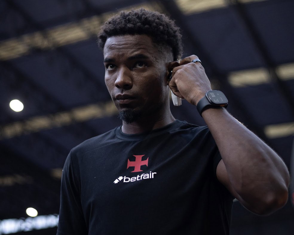 Homem negro com camiseta do Vasco, ajustando fone de ouvido no estádio.