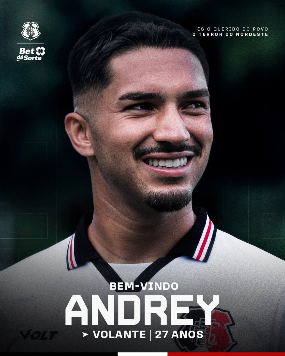Texto alt: Fotografia de um homem sorridente com cabelo escuro, barba por fazer, usando camisa de time de futebol, com fundo desfocado escuro e verde.