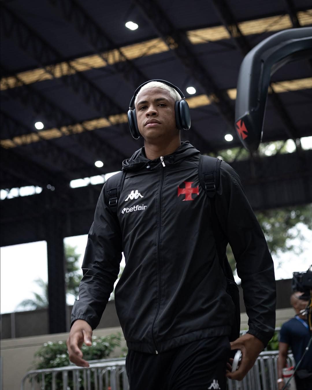 Atleta do Vasco com fones de ouvido, mochila e jaqueta preta, saindo do estádio em dia de jogo ou treino, com foco na força e determinação.