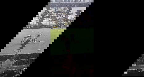 Jogadores do Vasco treinando na academia, com foco no campo de futebol cheio de marcas de treino. Torcedores podem ver a preparação do time.