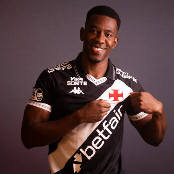 Jovem jogador do Vasco vestindo uniforme preto e branco com logos da equipe e patrocinadores, segurando a camisa e sorrindo para a câmera.