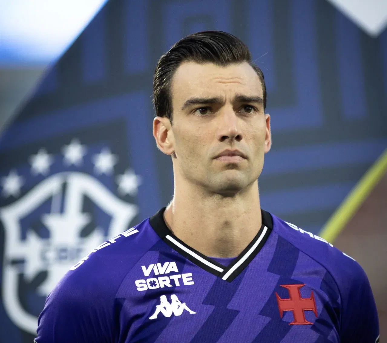 54803238641_1ff8583370_o Jogador do Vasco em uniforme azul escuro, fundo com brasão do clube Vasco da Gama.