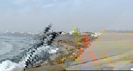 Jovem com uniforme do Vasco na praia de Santos, sorrindo e apontando para o mar, com cidade ao fundo e céu claro ao amanhecer ou entardecer.
